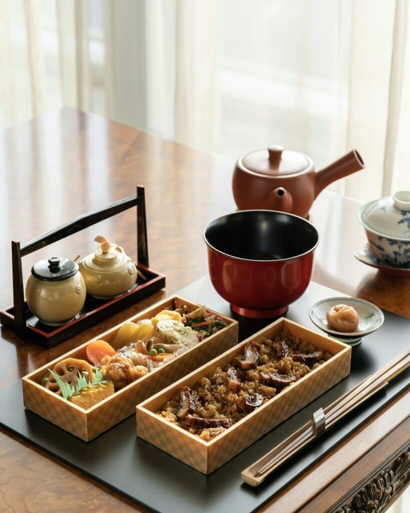 Two wooden lunch boxes packed with rice and cooked vegetables on a tray beside a teapot and a red bowl