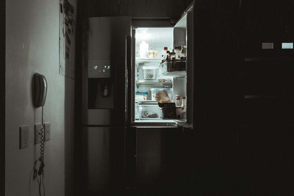 An open French-door refrigerator glowing in a dark kitchen, its shelves holding containers and bottles
