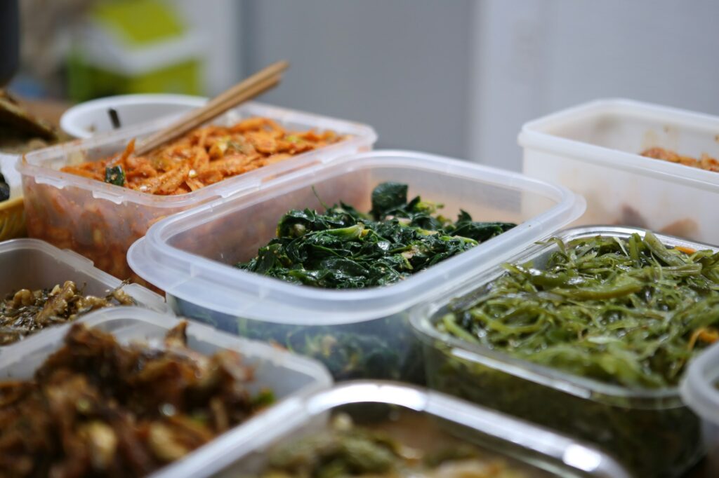 Clear plastic food containers filled with cooked greens, carrot salad and other prepared dishes on a counter