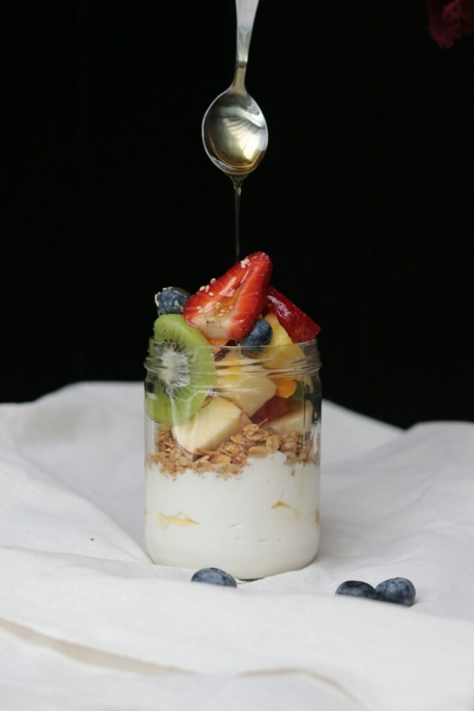 A spoon drizzling honey over a jar of yogurt, granola and cut fruit