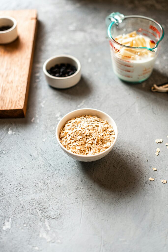 A bowl of rolled oats, a glass measuring cup of milk and a small dish of dark berries on a grey table