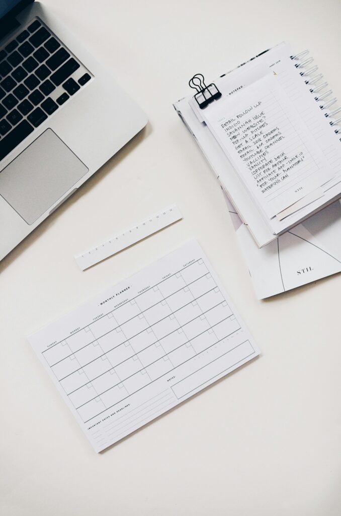 A laptop next to a blank monthly planner sheet and a clipped notepad on a white desk