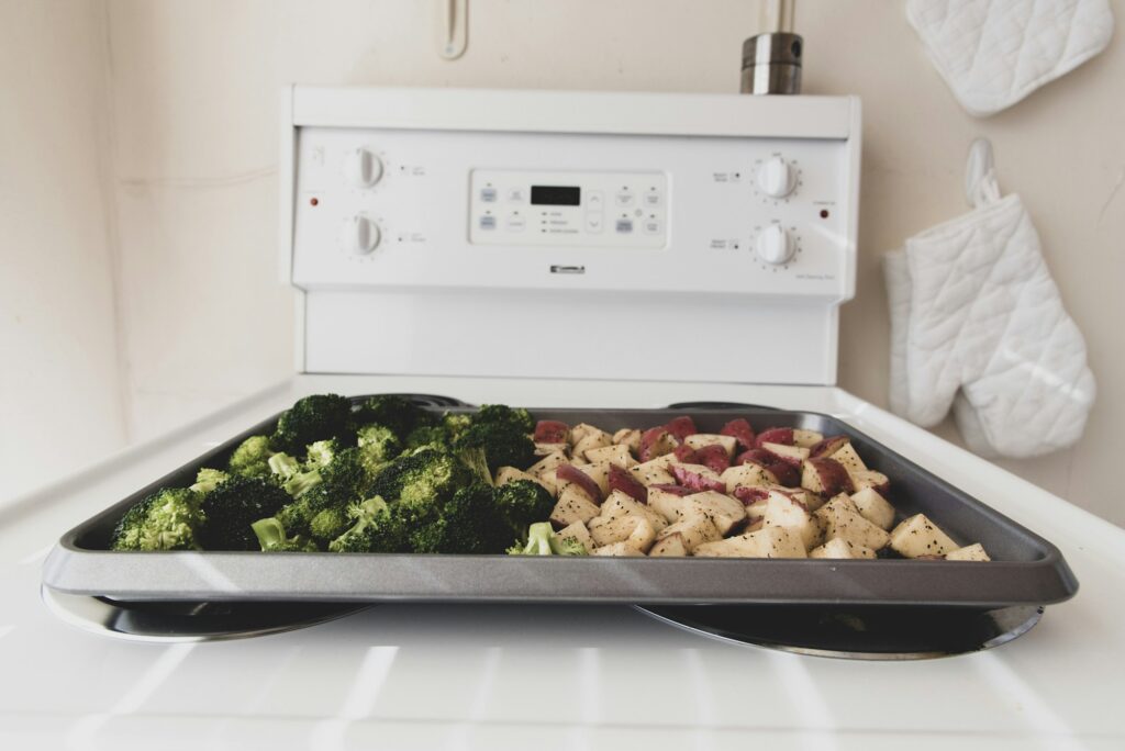A sheet pan of broccoli and diced potatoes going into an open oven, oven mitts hanging on the wall